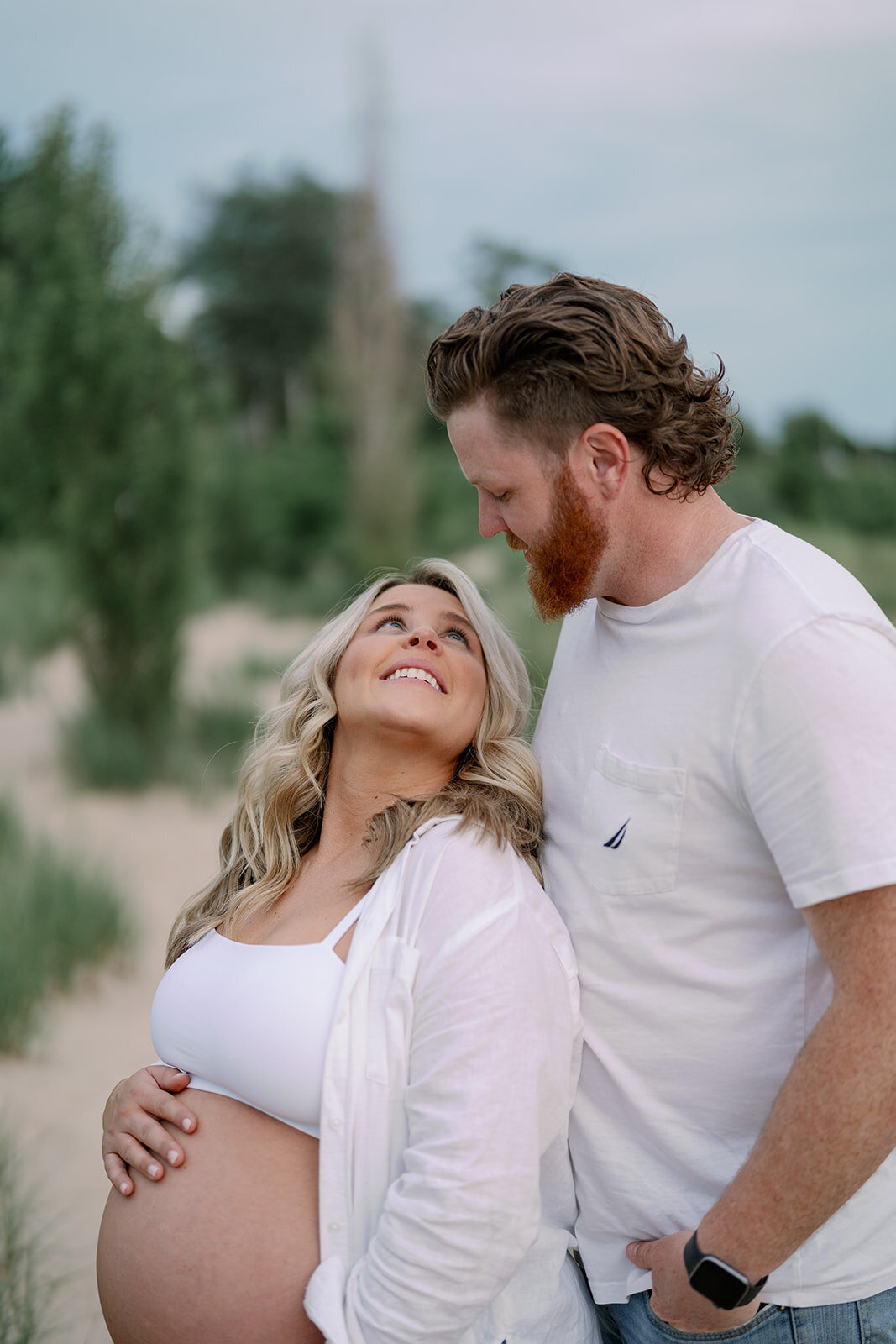 Expecting couple smiling at each other in the beach grass during their South Haven North Beach maternity session.