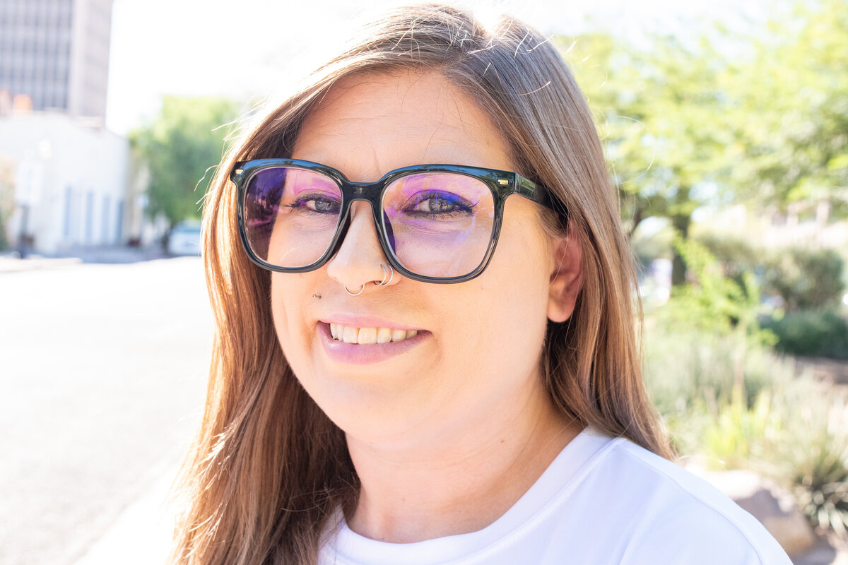 Woman smiling outdoors in bright sunlight, wearing large glasses and facial piercings, photographed by Vyrl Photo, showcasing Tucson brand photography.