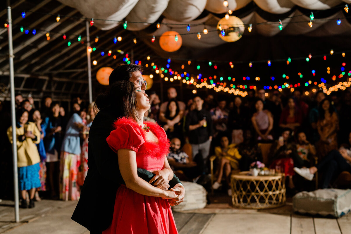 The bride, changed into her red reception dress, dances with the groom in a tight embrace under the colorful lights in the reception in the greenhouse at Dos Pueblos Orchid Farm.