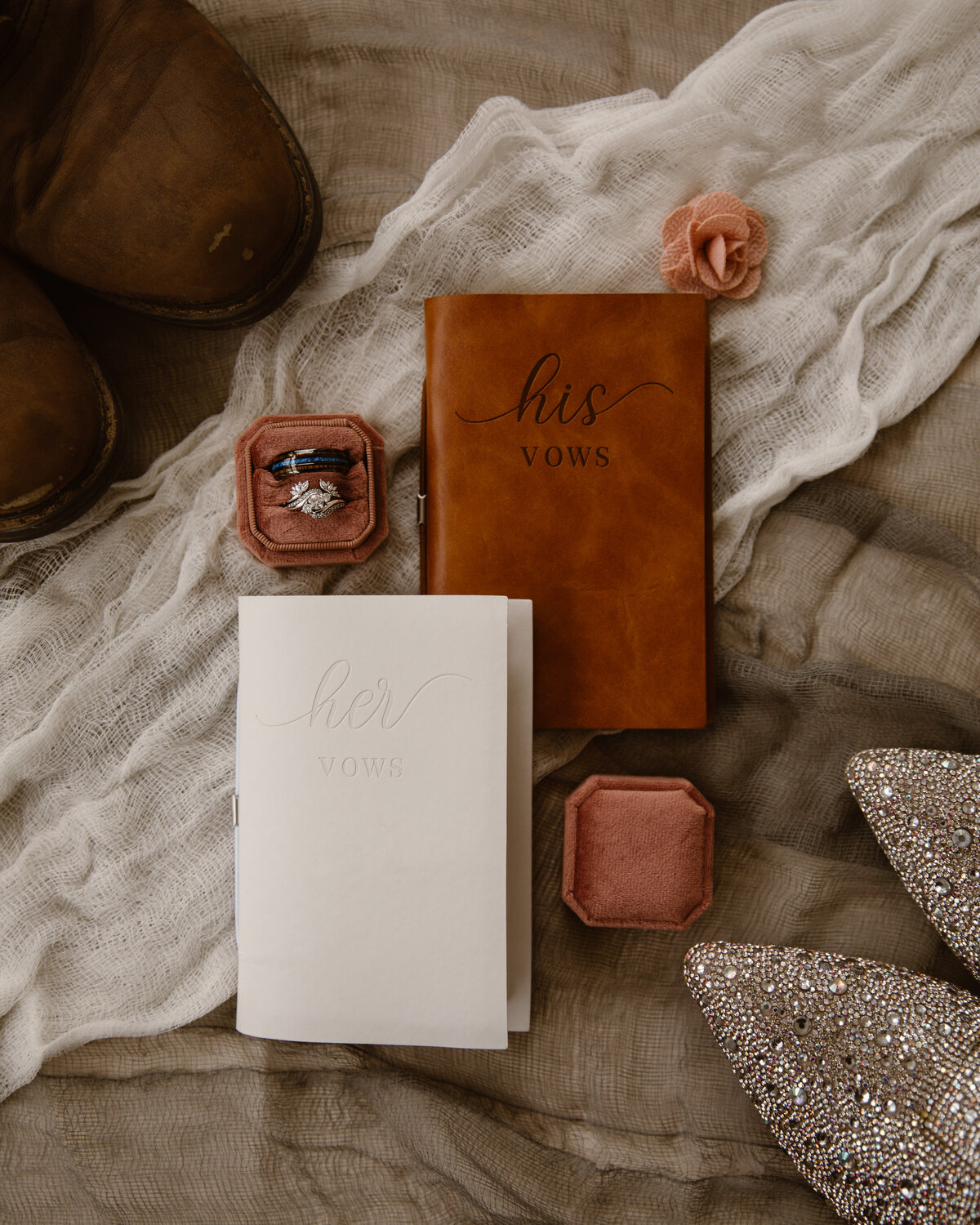 A flat lay of rustic wedding details including “his” and “her” vow books, sparkling bridal shoes, blush velvet ring boxes with wedding bands, and a soft textured fabric background — perfect for a Colorado wedding day story.