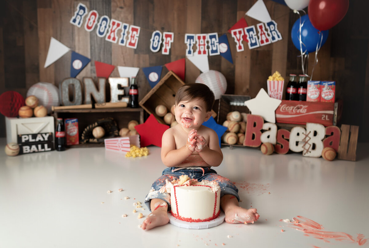 Baseball themed cake smash at West Palm Beach and Boynton Beach newborn and cake smash photographer. Baby boy is wearing blue jeans and no shirt sitting behind a baseball themed cake. There is cake all over his pants and hands. He is smiling at the camera. In the background, there are vintage baseballs, soda pop bottles, and signage.