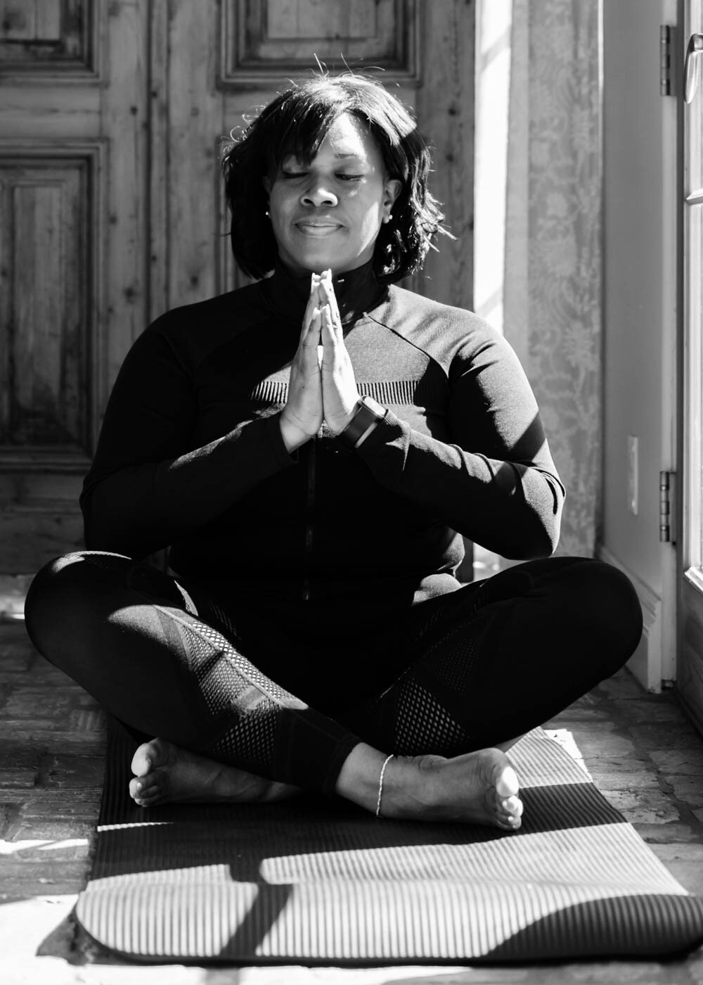 A black and white portrait of a woman sitting on the floor next to a window with light on a yoga pose