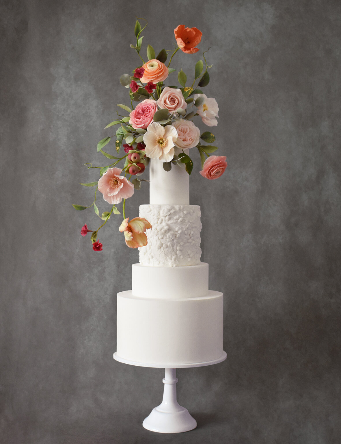 A white four tiered wedding cake with bas relief detail and Dutch Master style sugar flower arrangement