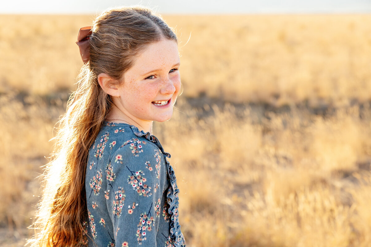 A young girl with a brown bow in her hair smiles softly while looking into the distance, standing in a golden wheat field.