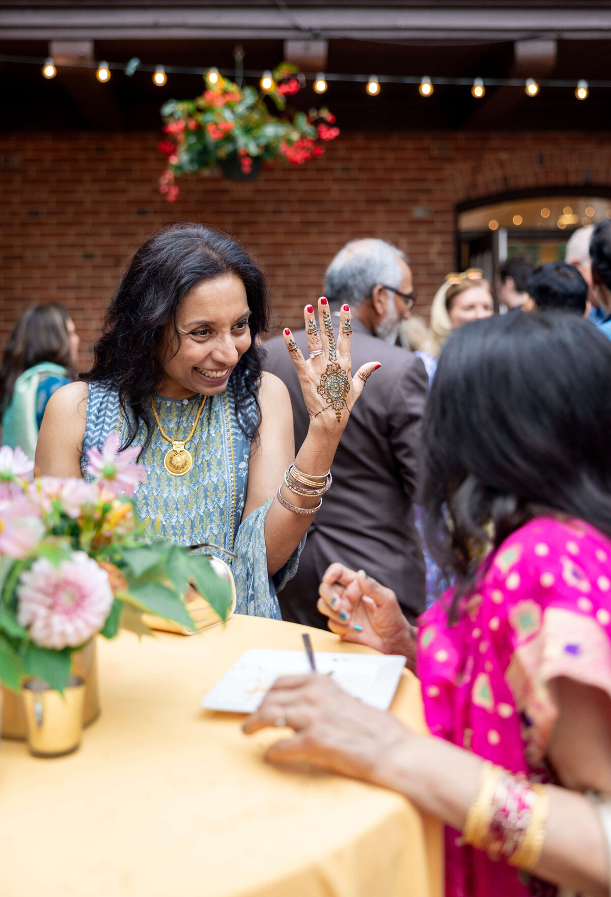 A woman in a patterned dress proudly displays intricate henna on her hand at a gathering. She is standing at a table with flowers, engaging with another person in a pink saree. The background shows people mingling outdoors.