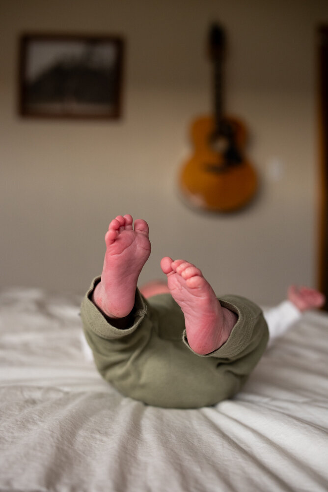 Close-up of a newborn baby's feet sticking up in the air while lying on a bed. The baby is wearing olive green pants, and the soft lighting creates a warm, cozy atmosphere. A blurred acoustic guitar and framed photo are visible in the background