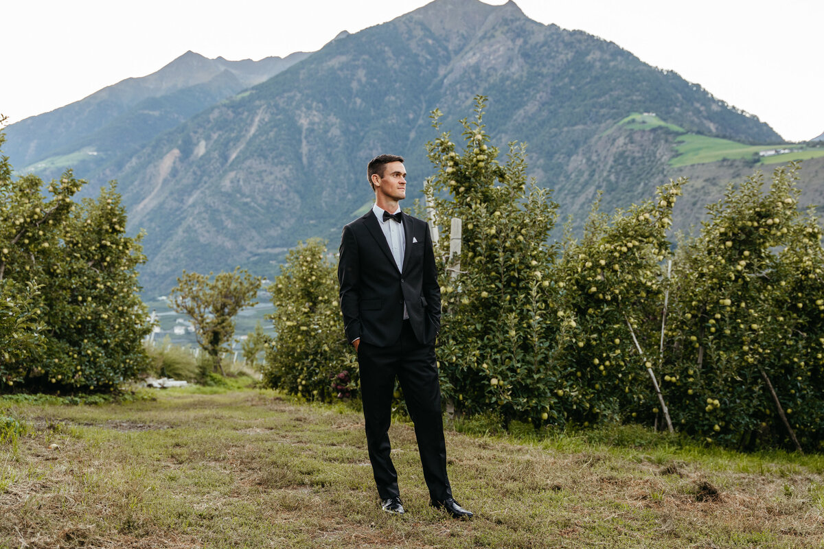 Groom standing in orchard with mountains behind him