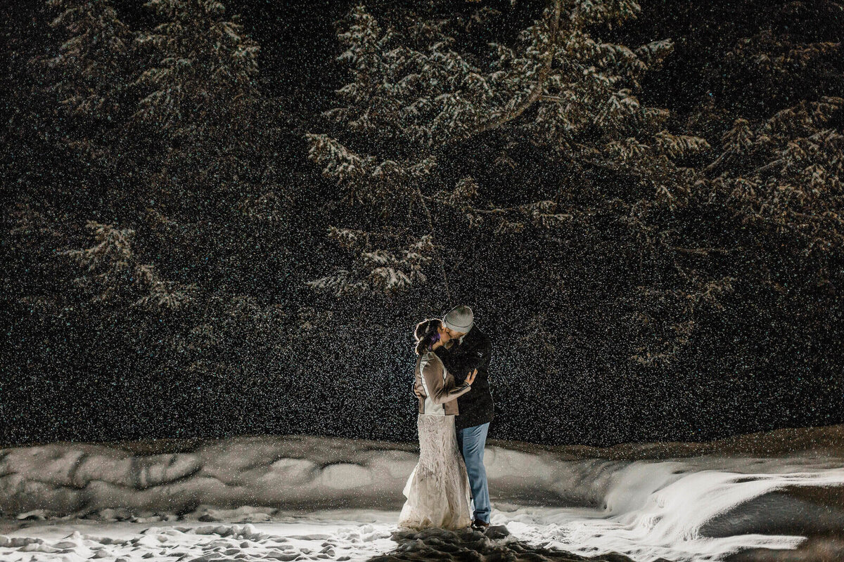 A bride and groom kiss while standing near a snowbank and the snow falls heavily during their winter elopement in Washington