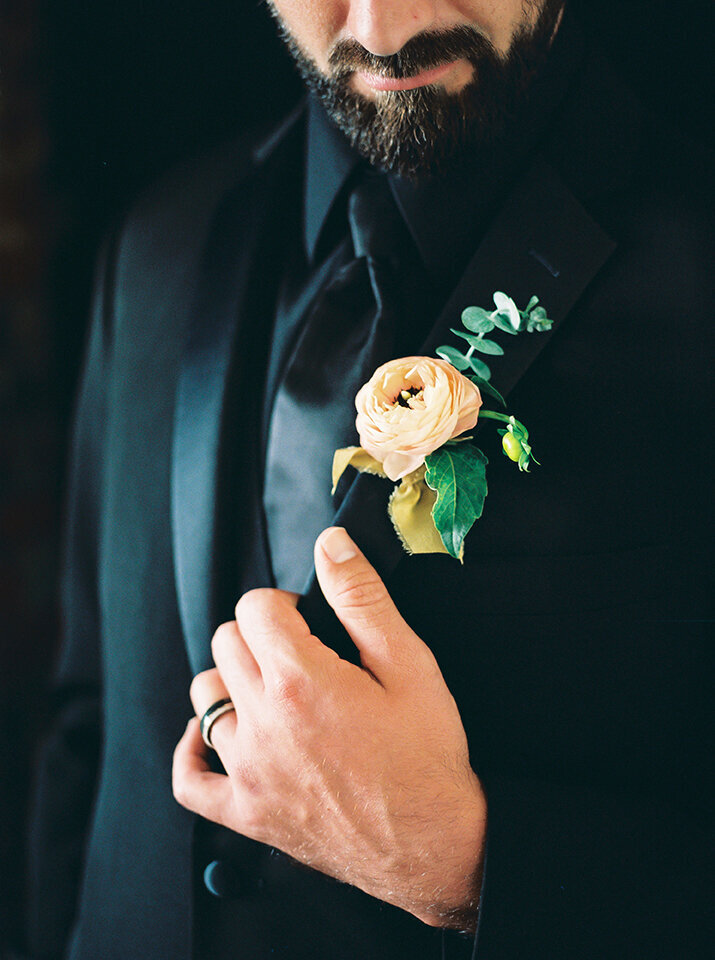 A close-up of the groom wearing a black tuxedo with a flower tucked in front pocket.