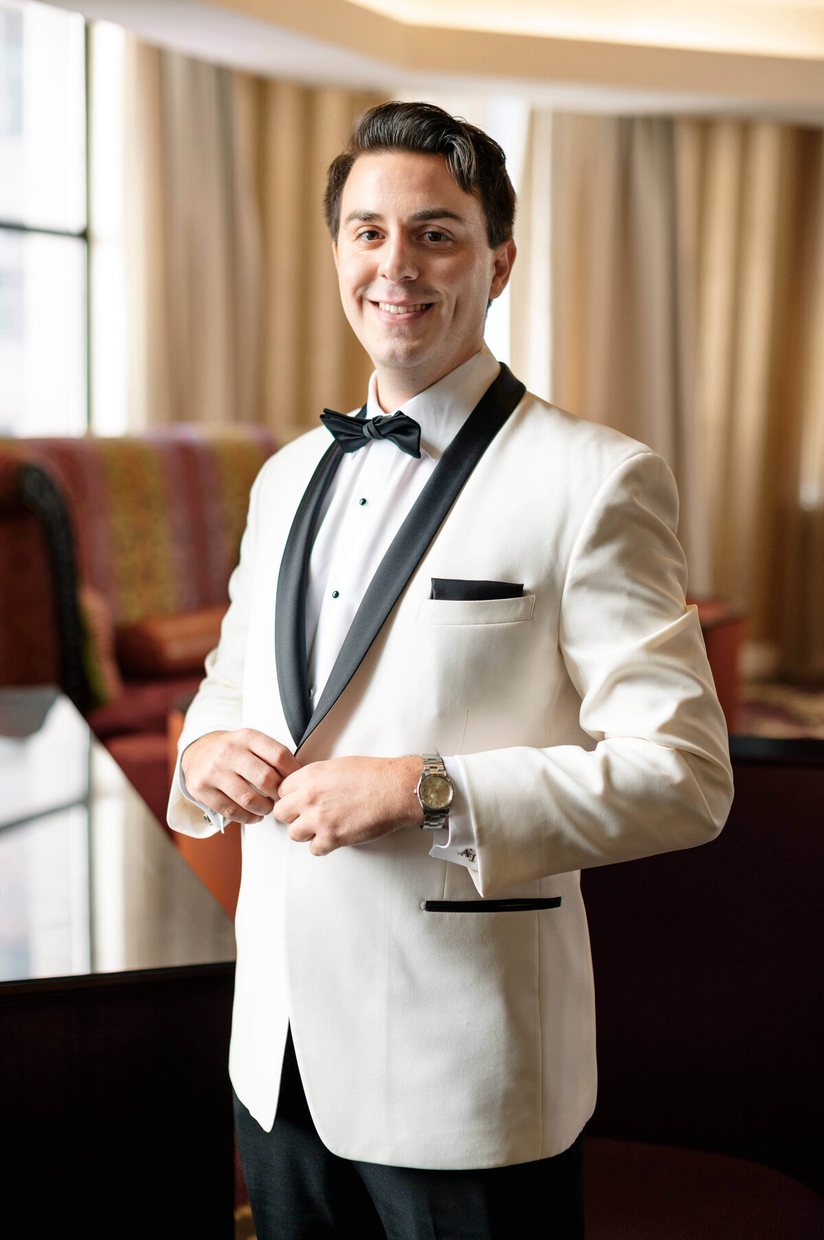 A man stands indoors wearing a white tuxedo with black accents and a bow tie, smiling while buttoning his jacket. The background features a softly lit room with draped curtains and furniture.