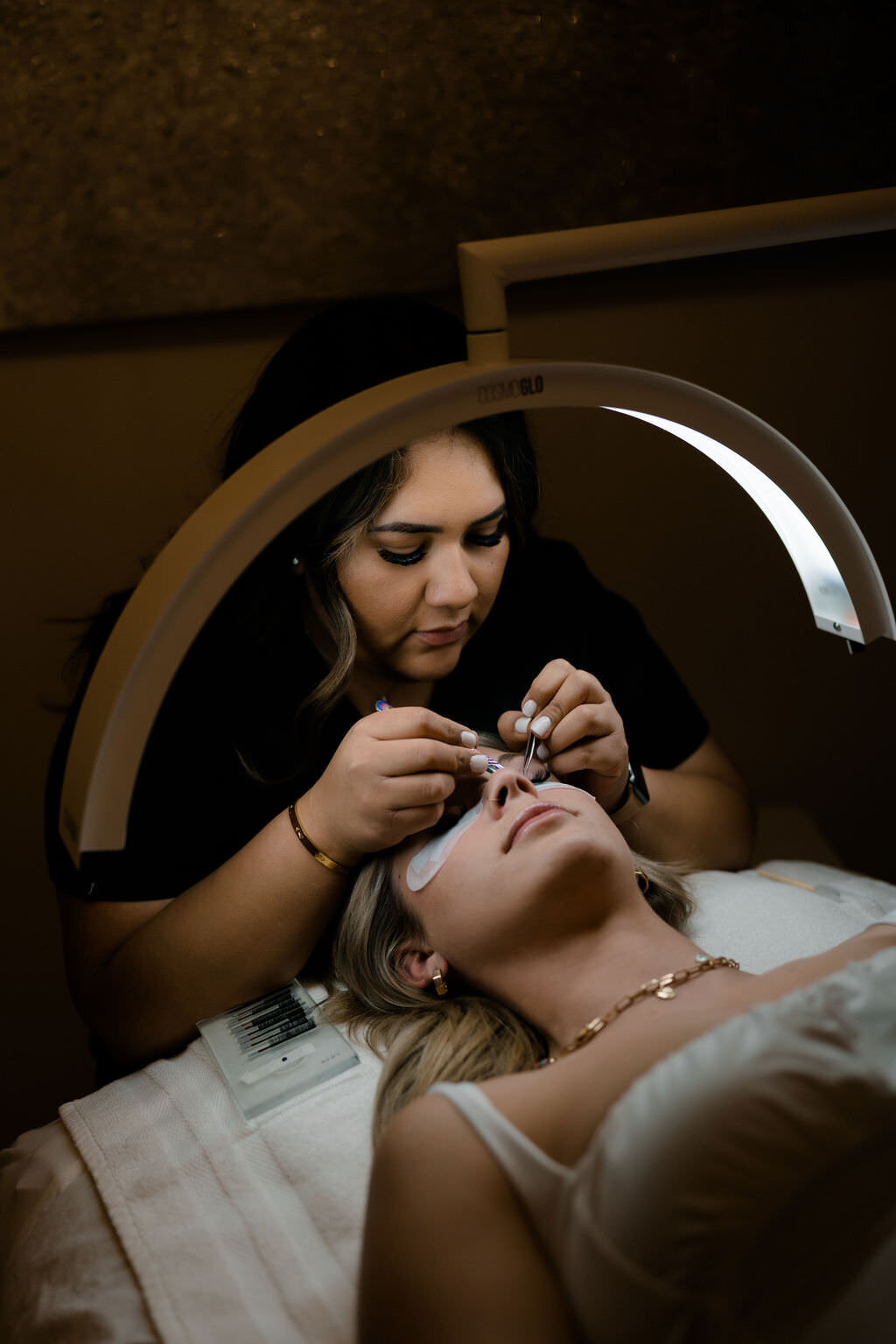 woman placing eyelashes on a customers eyes with a half circle light over them