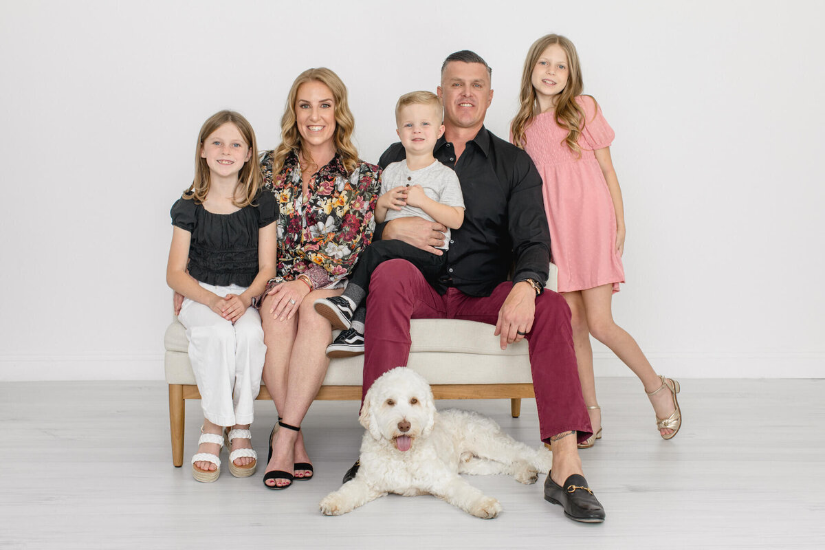 A family of five with dirty blonde hair, wearing shades of black and pink sitting on a white bench with their white, fluffy dog, smiling at the camera in a white studio in Tustin.