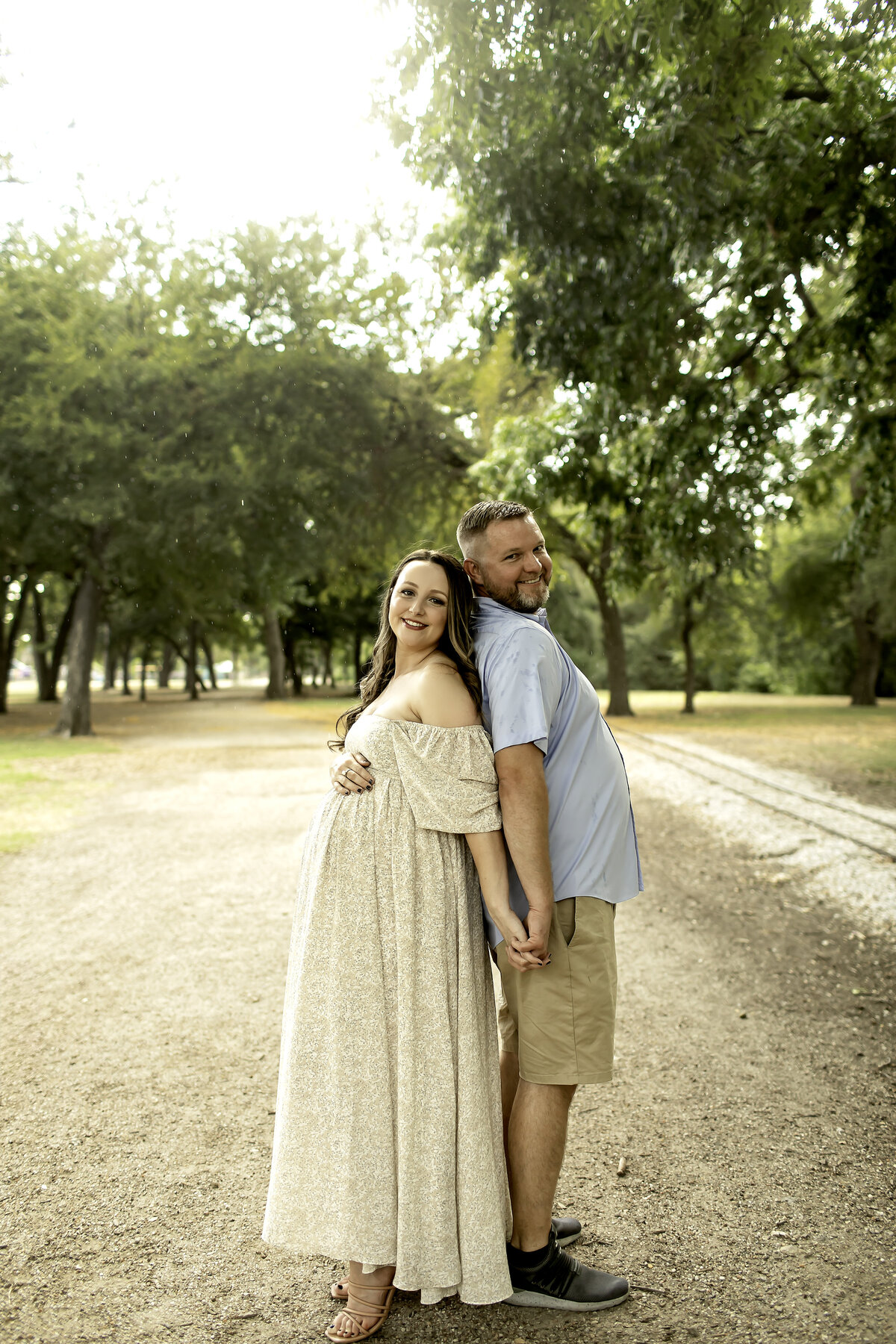 A joyful maternity photo of mom and dad laughing together in the beautiful greenery of Trinity Park, with mom lovingly cradling her belly, captured by Chunky Monkey Photography.