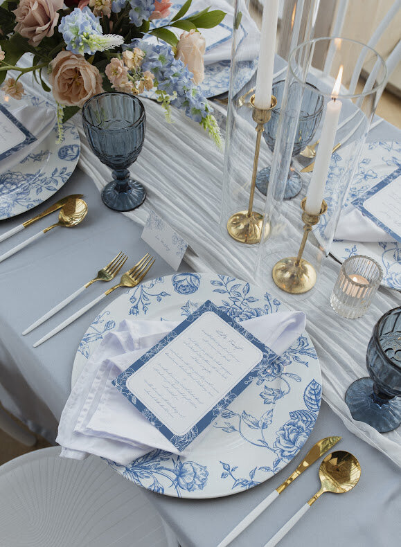 A close-up of an elegant guest table at a National Arts Centre wedding, featuring dusty blue linens, French toile floral patterned blue charger plates, and stylish blue floral menus layered on top. The table is adorned with gold and white cutlery, tapered candles, vibrant flowers, and sophisticated blue goblets.