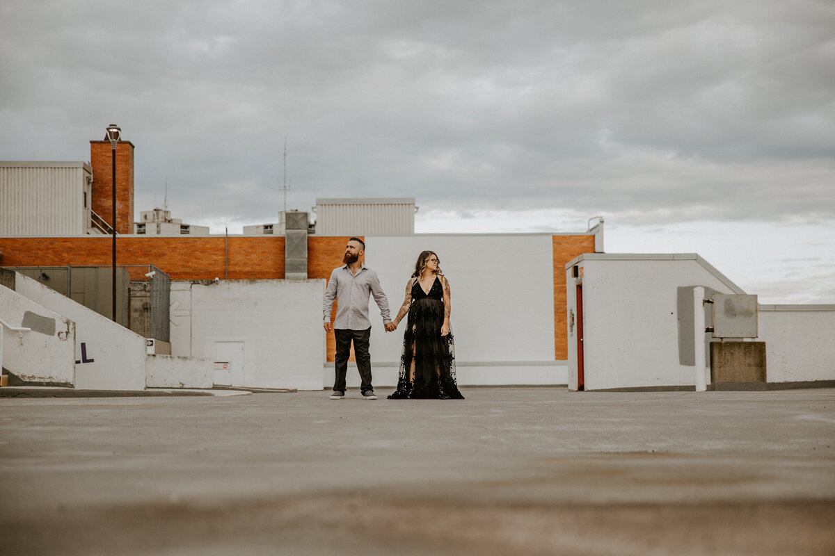 Portrait at London, Ontario engagement session. The man and woman are standing and holding hands in the distance, both looking off camera in opposite directions. Photo captured at the top of a downtown London, Ontario parking garage.