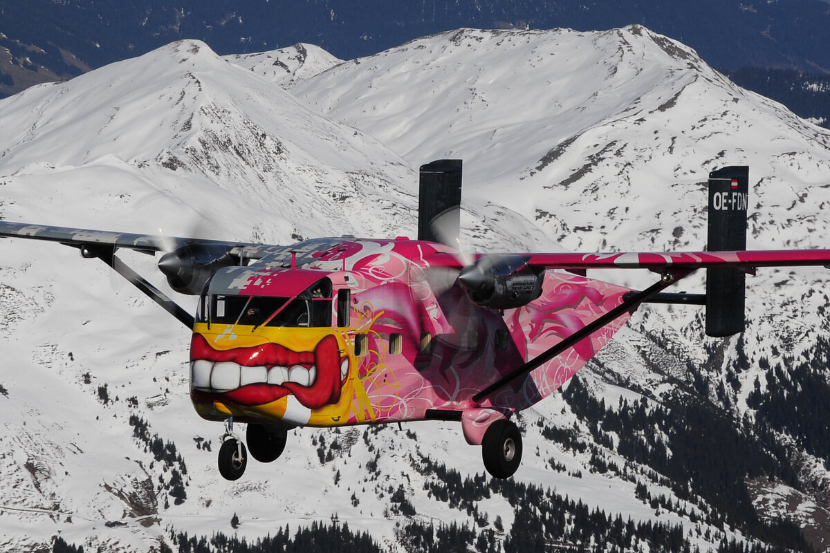 Die Pink Skyvan fliegt über die verschneiten Alpen. Zu sehen ist das Flugzeug im Flug. Die bunte und auffällige Lackierung sticht heraus