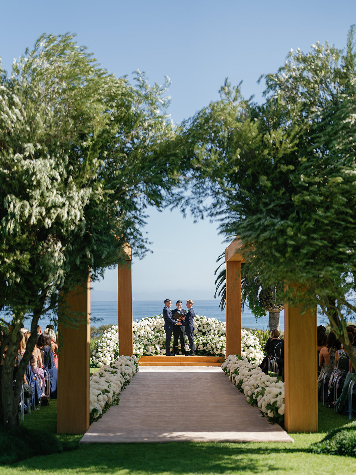 Luxurious wedding ceremony at The Ritz Carlton Bacara with lush white flowers, olive trees, carpeted aisle and wood shade structures