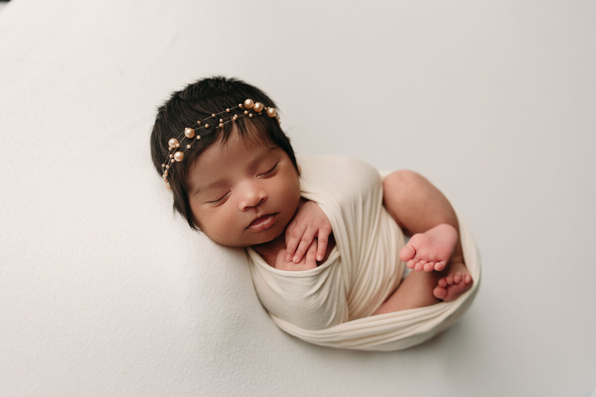 Baby newborn girl with thick brown hair wearing a pearl headband lying on back swaddled in white laying on white backdrop