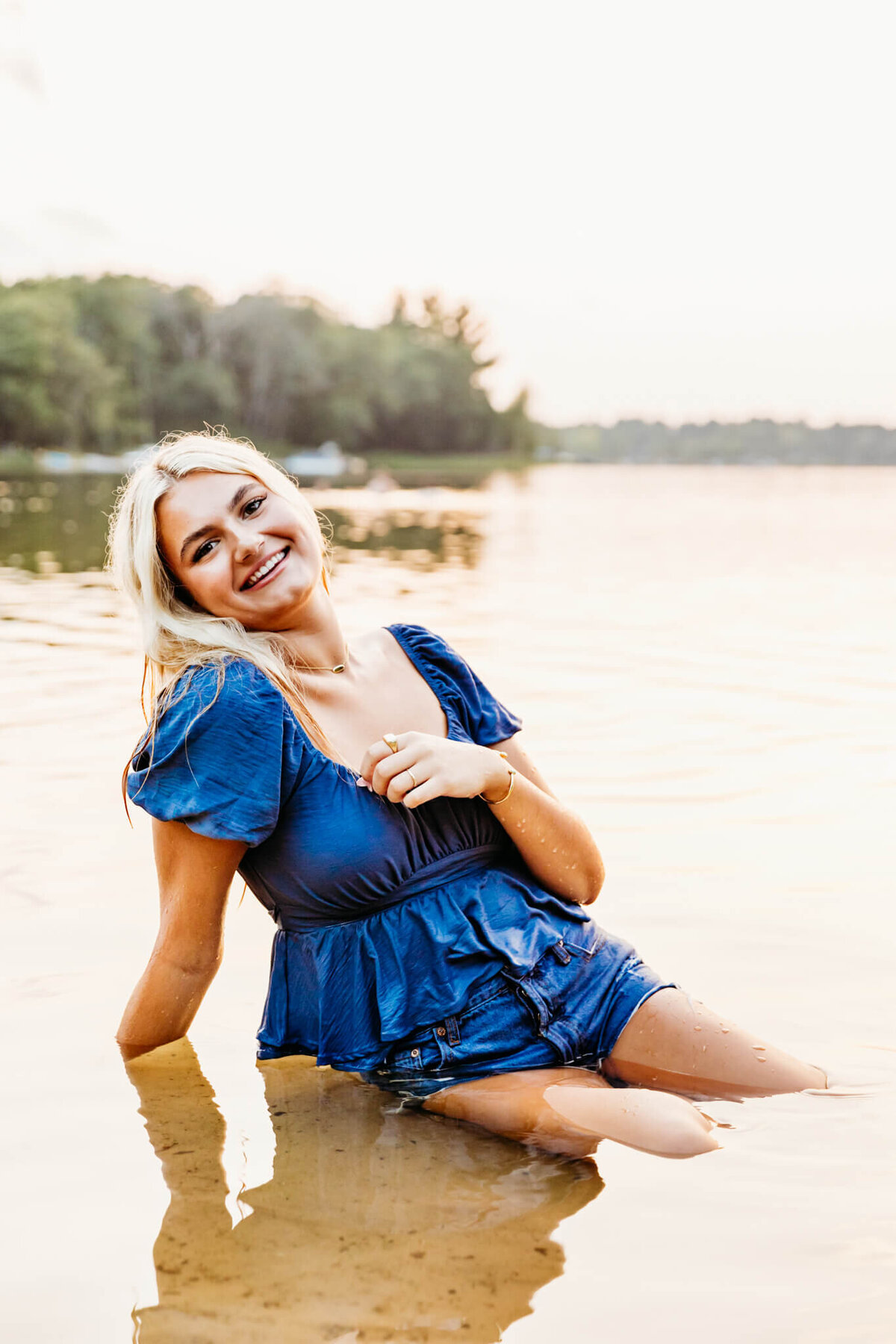 teenage girl in a blue top and jean shorts sitting in a shallow lake near Neenah and laughing 