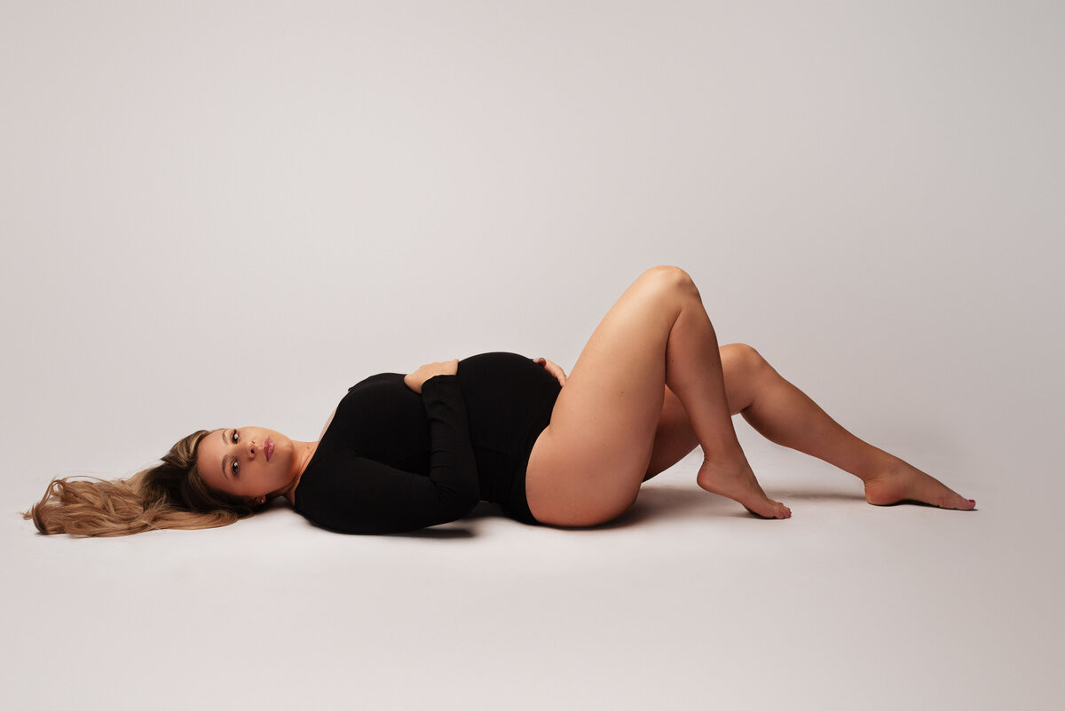 blonde mom to be laying on floor looking at the camera holding her baby bump smiling at the camera in black bodysuit over white backdrop in studio