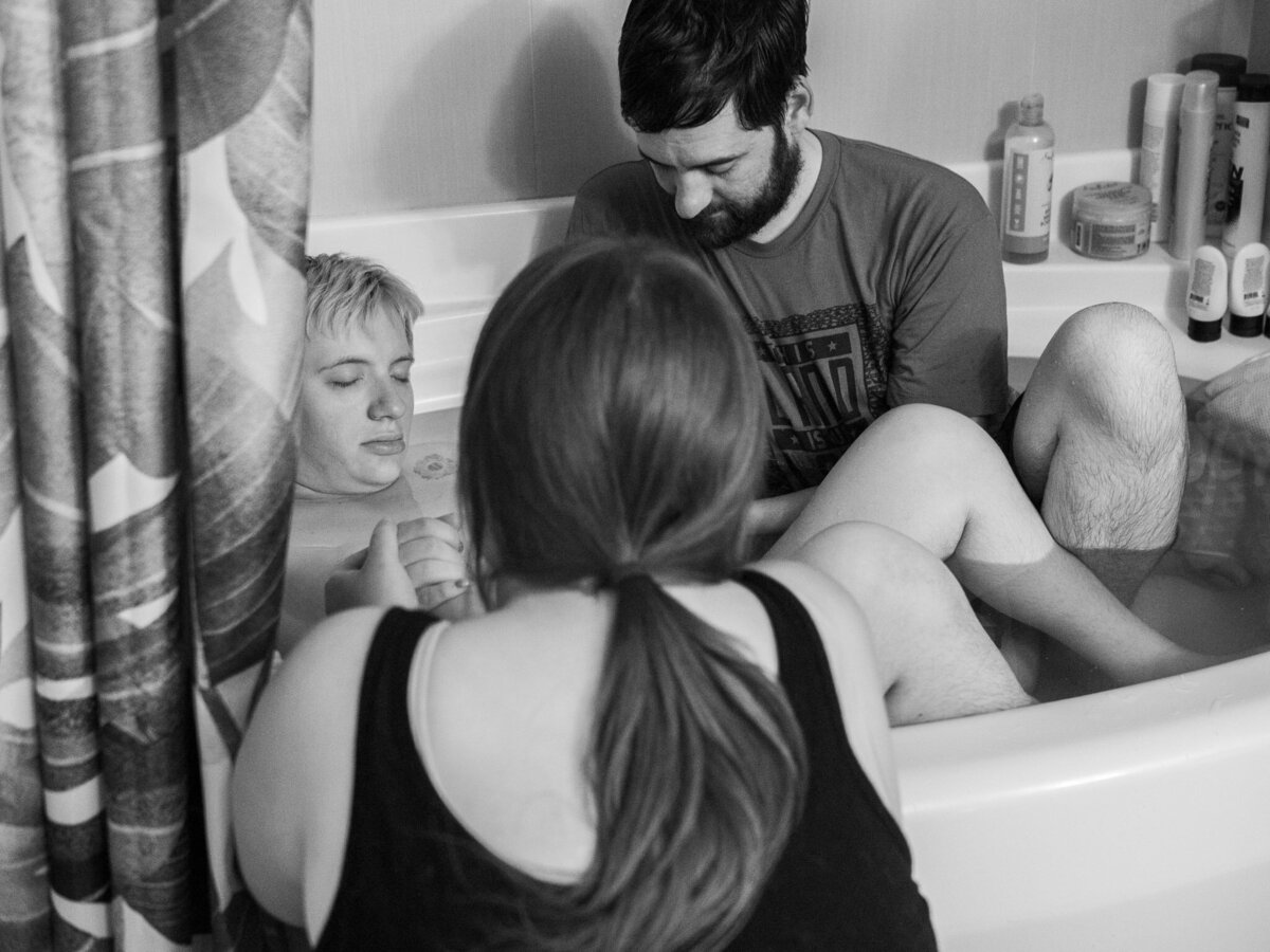 A pregnant person and their husband is laboring in a tub at a home birth with their doula sitting outside of the tub supporting in Shoreline, WA.