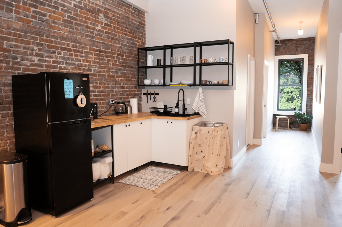 Kitchen for collective members with black shelves holding glasses over a white and wood kitchenette. A large, black refrigerator stands against an exposed brick wall.