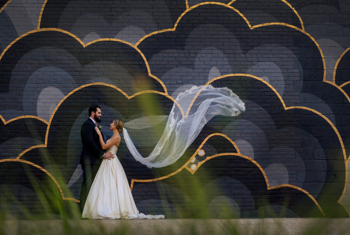 The bride's veil flies as she and her husband hold each other in front of a colorful mural.