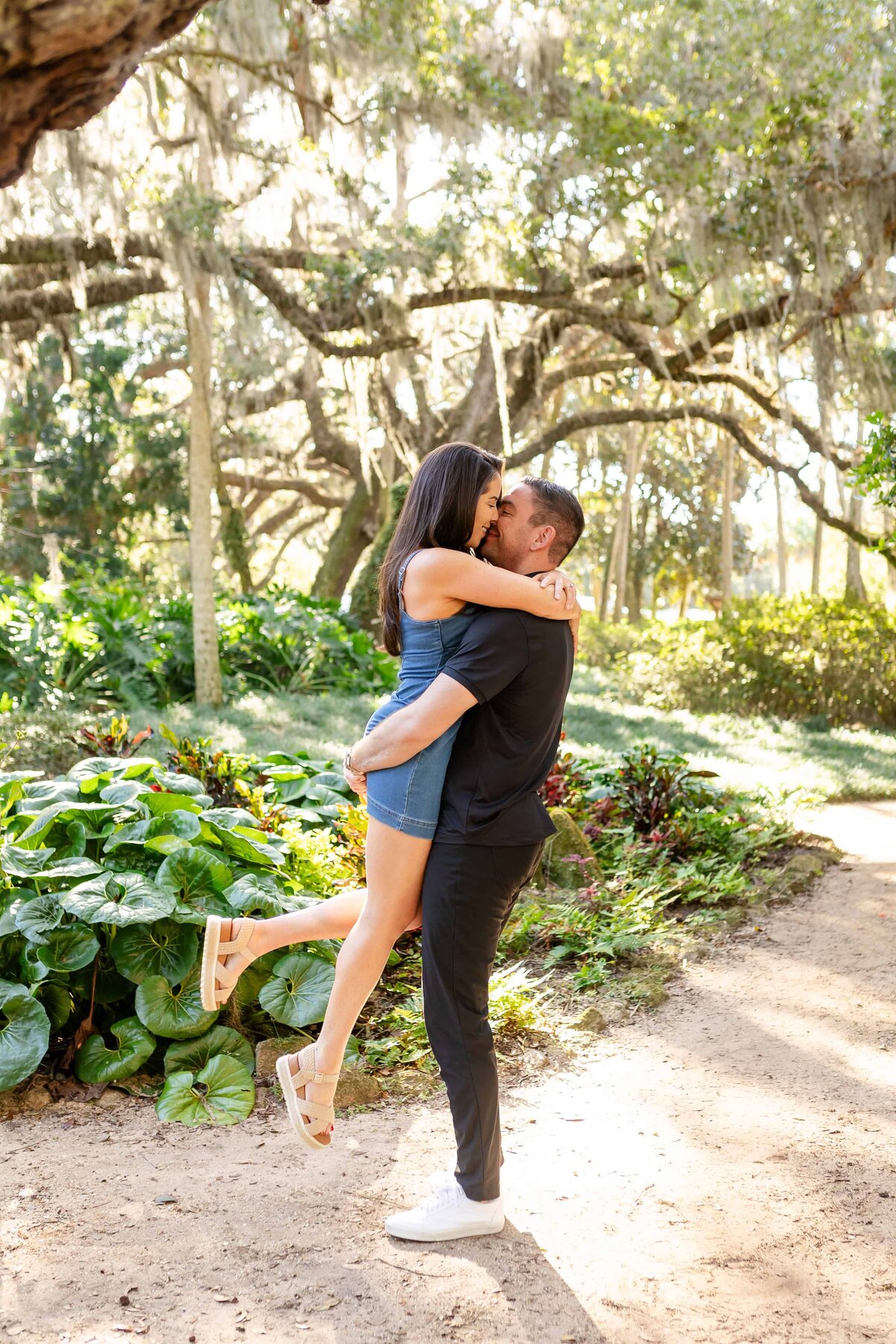  A joyful couple shares a kiss as the groom-to-be lifts his bride-to-be in a romantic embrace surrounded by lush greenery and sunlight in Jacksonville, Florida. She wears a denim mini dress and sandals while wrapping her arms around his neck, creating a spontaneous and loving moment. Captured by Heartful Films, a Jacksonville engagement photographer and videographer known for fun, energetic, and authentic storytelling in natural settings.