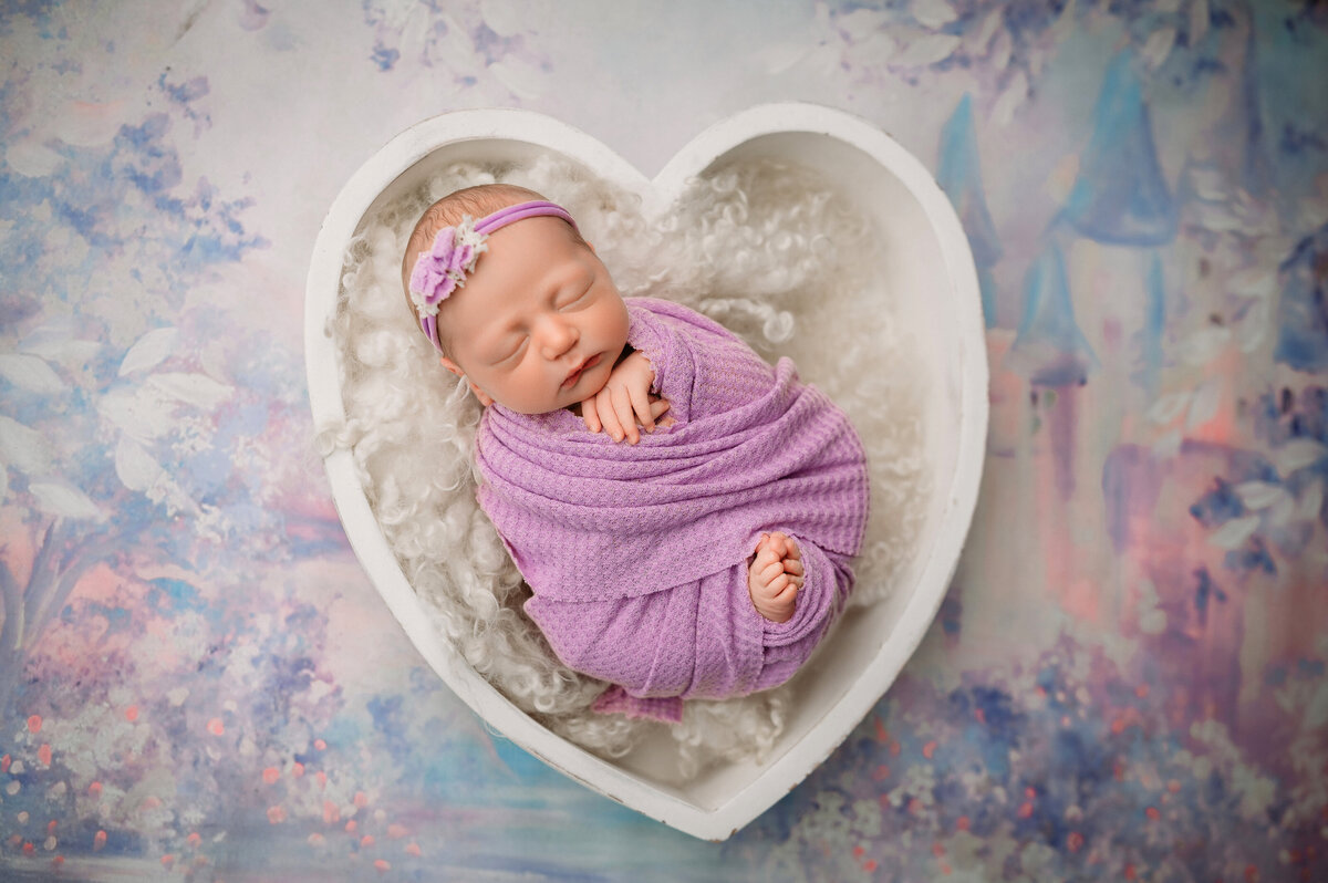 newborn baby girl wrapped in  purple swaddle with feet and hands poking out and posed in a white heart bowl prop with a whimsical castle backdrop with purple blues, and pinks