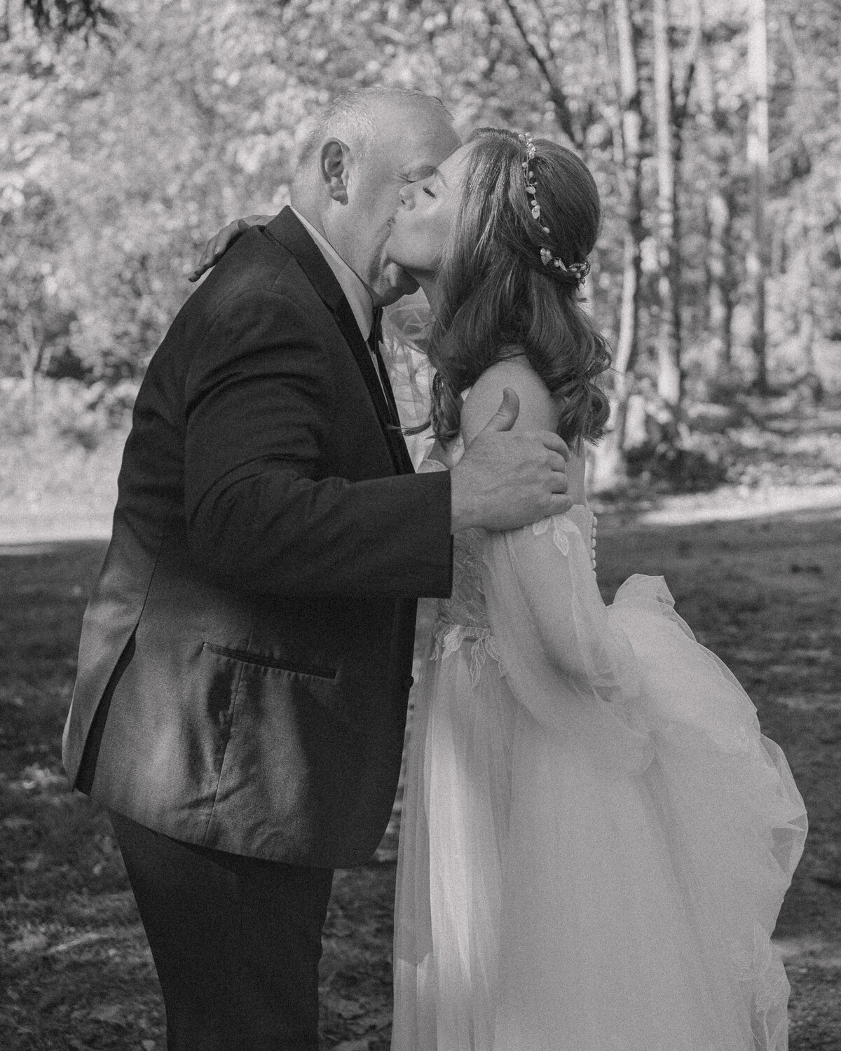 Heartwarming black and white photo of a father sharing a tender kiss with his daughter, the bride, on her wedding day. The bride, in a delicate lace gown, embraces her father amidst a serene, wooded backdrop, capturing a candid, emotional moment in a documentary style.