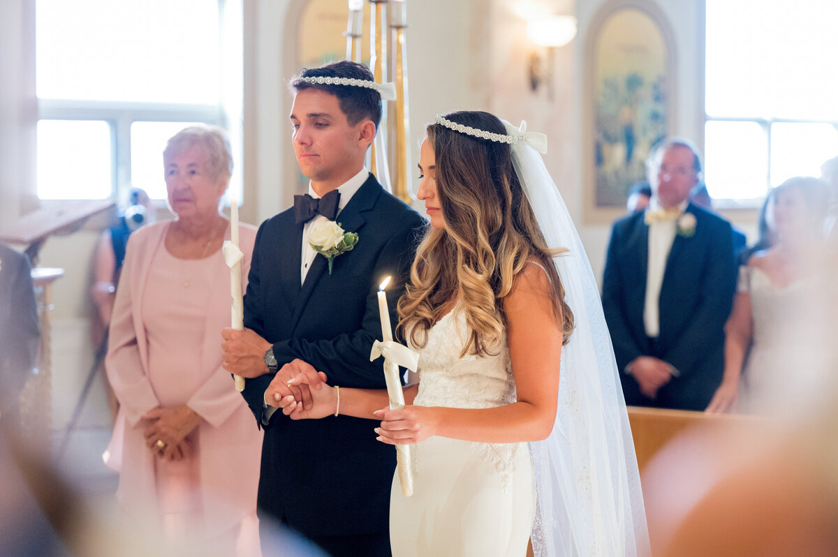 bride and groom at greek orthodox wedding ceremony wearing crowns and holding candles in religious symbol