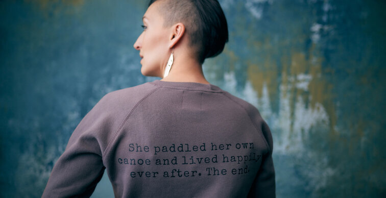 Tarzan has her back to the camera and looks off to the side, the back of her sweater reads “She paddled her own canoe and lived happily ever after. The end.”