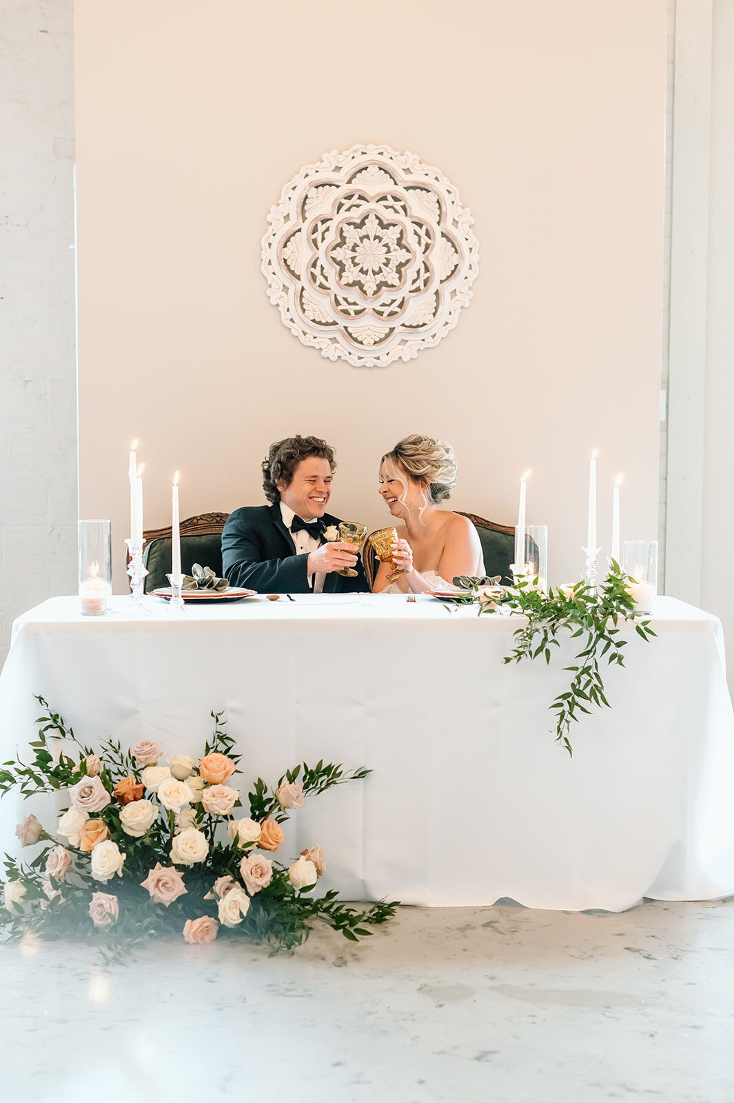A bride and groom clink glasses in a romantic, candlelit setting, celebrating their new life together with smiles and joy at their beautifully decorated reception table.