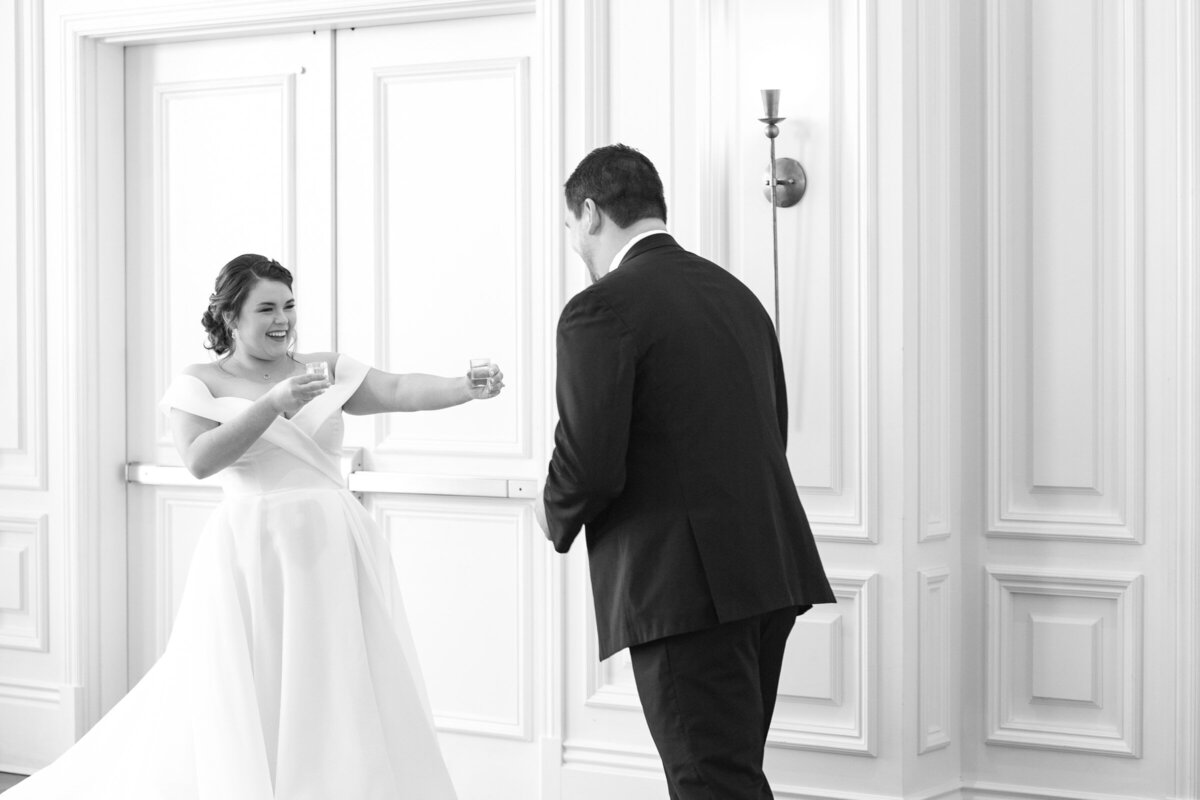 black and white photo of the bride offering her groom a shot of tequila during their first look in the Governor’s Room at The Adolphus in Dallas, capturing a playful and candid wedding moment.