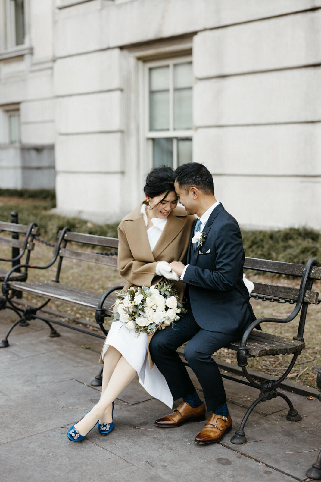 The bride and the groom are sitting closely together, touching heads, on a bench outside of a building.