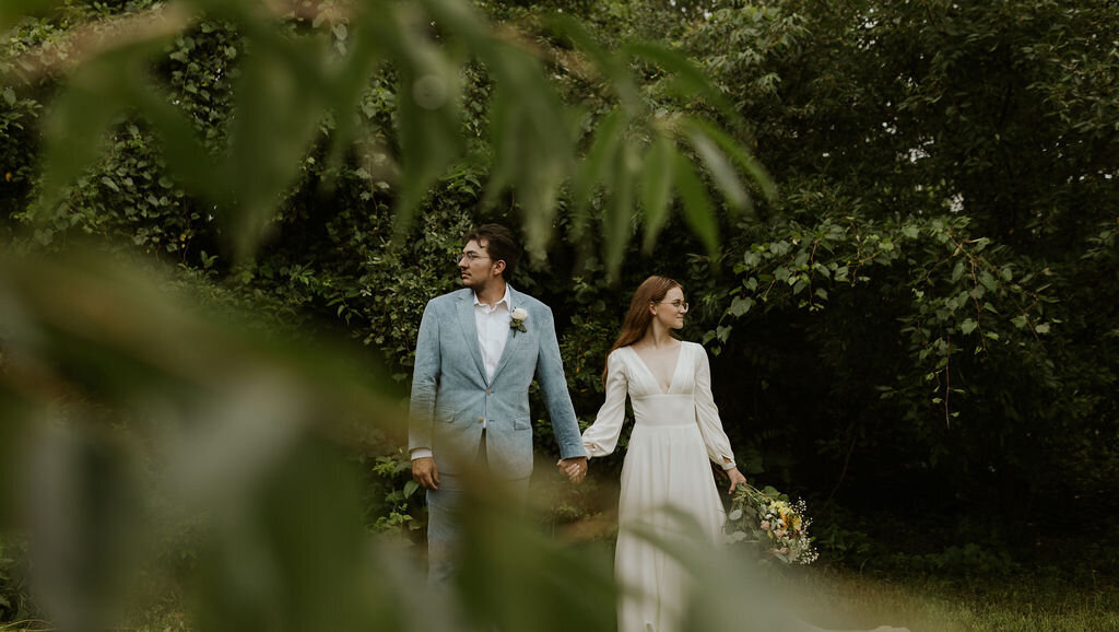 Bride and groom facing the camera holding hands while looking apart from eachother. They are standing in front of deep green trees