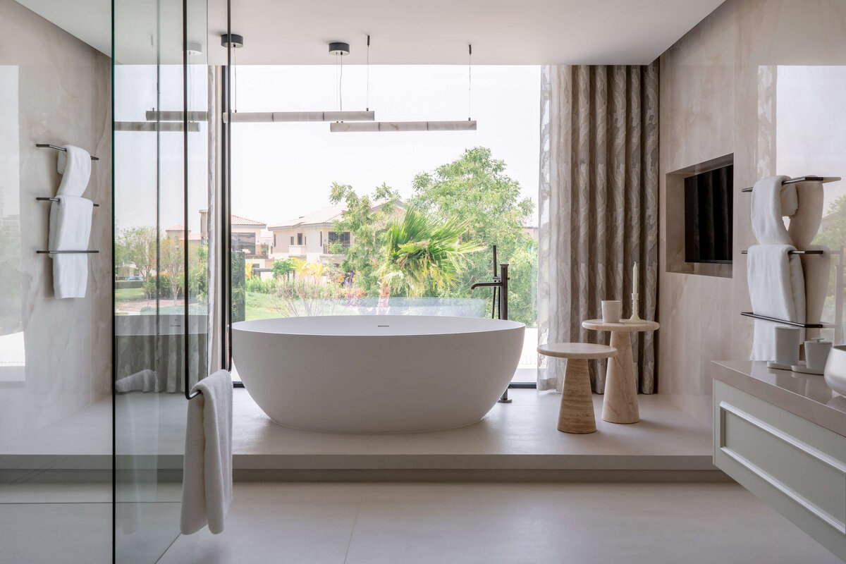 Large soaking tub in the primary bathroom. The wall behind the tub is entirely a window, looking out to a lush green scene. To the right of the tub is a recessed television and two small, chic tables.