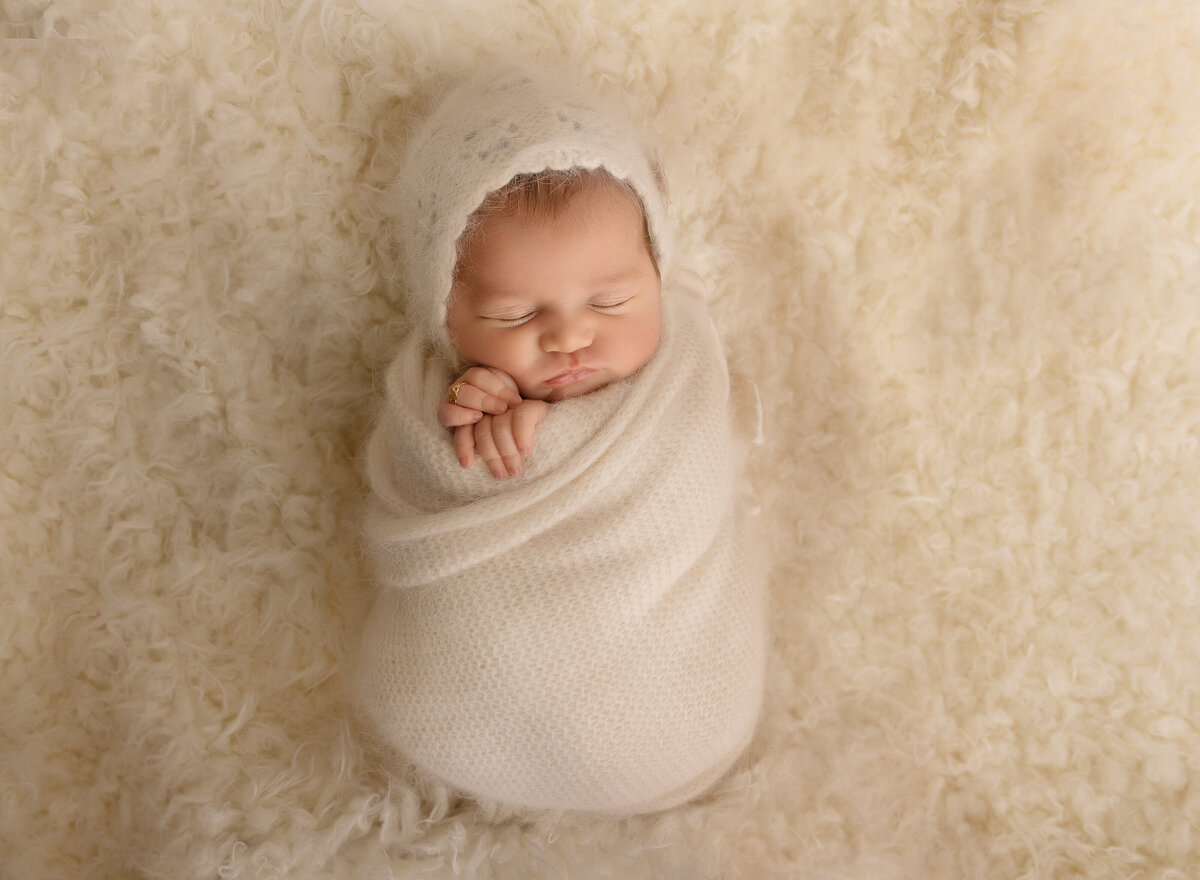 Baby in cream knit swaddle and bonnet is sleeping for their brooklyn newborn photoshoot. Baby's hands are peeking out of the swaddle. Captured by top Brooklyn newborn photographer Chaya Bornstein