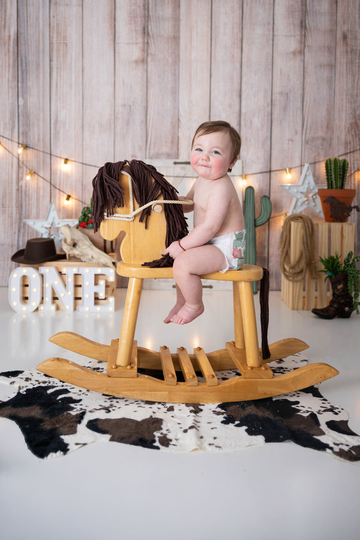 Baby wearing only a diaper, happily riding a wooden rocking horse with coordinating cowboy props in the background.