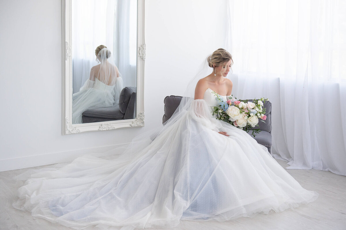 a romantic photo of a bride in a soft blue wedding dress sitting on a grey couch holding a bouquet with a creative mirror reflection behind her.  Captured by Ottawa wedding photographer JEMMAN Photography