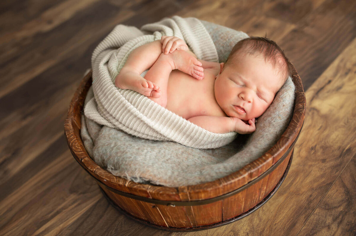 A newborn baby boy is wrapped in a white blanket and sleeps in a brown wood bucket
