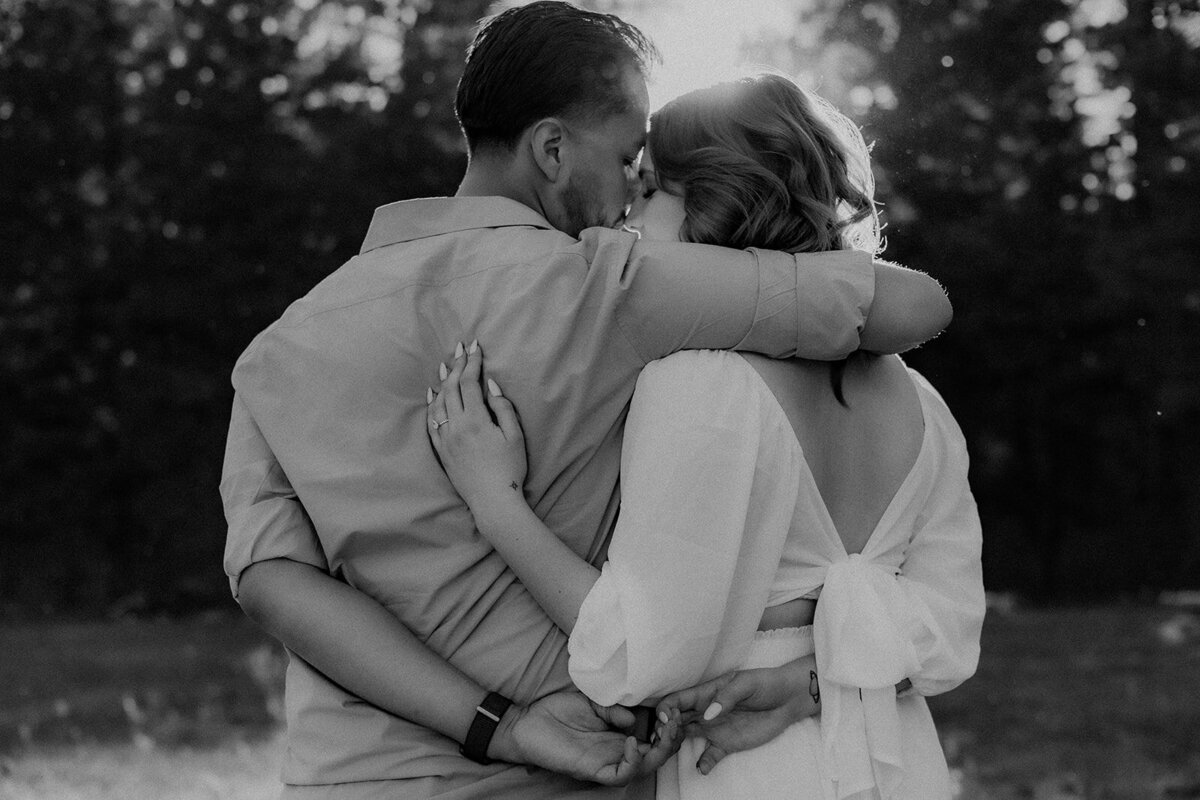 A monochrome image capturing the couple embracing from behind, their hands clasped together as sunlight streams through the trees.