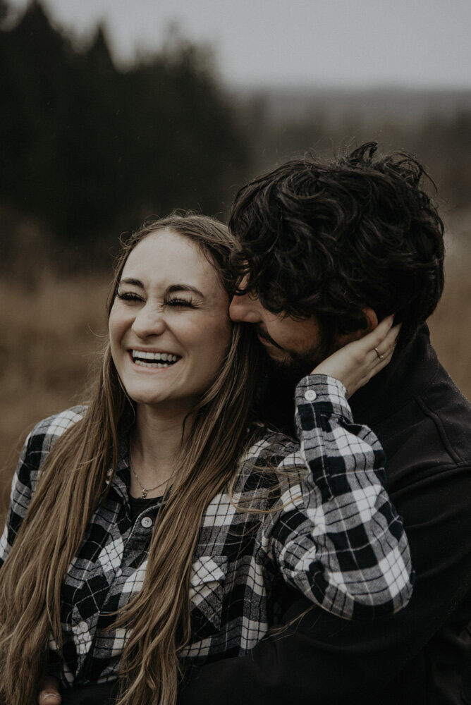 photo of a couple laughing together. The guy is behind the girl with his arms around her waist. Her left hand is reaching up to rest on his cheek while he nuzzles his nose against her temple.