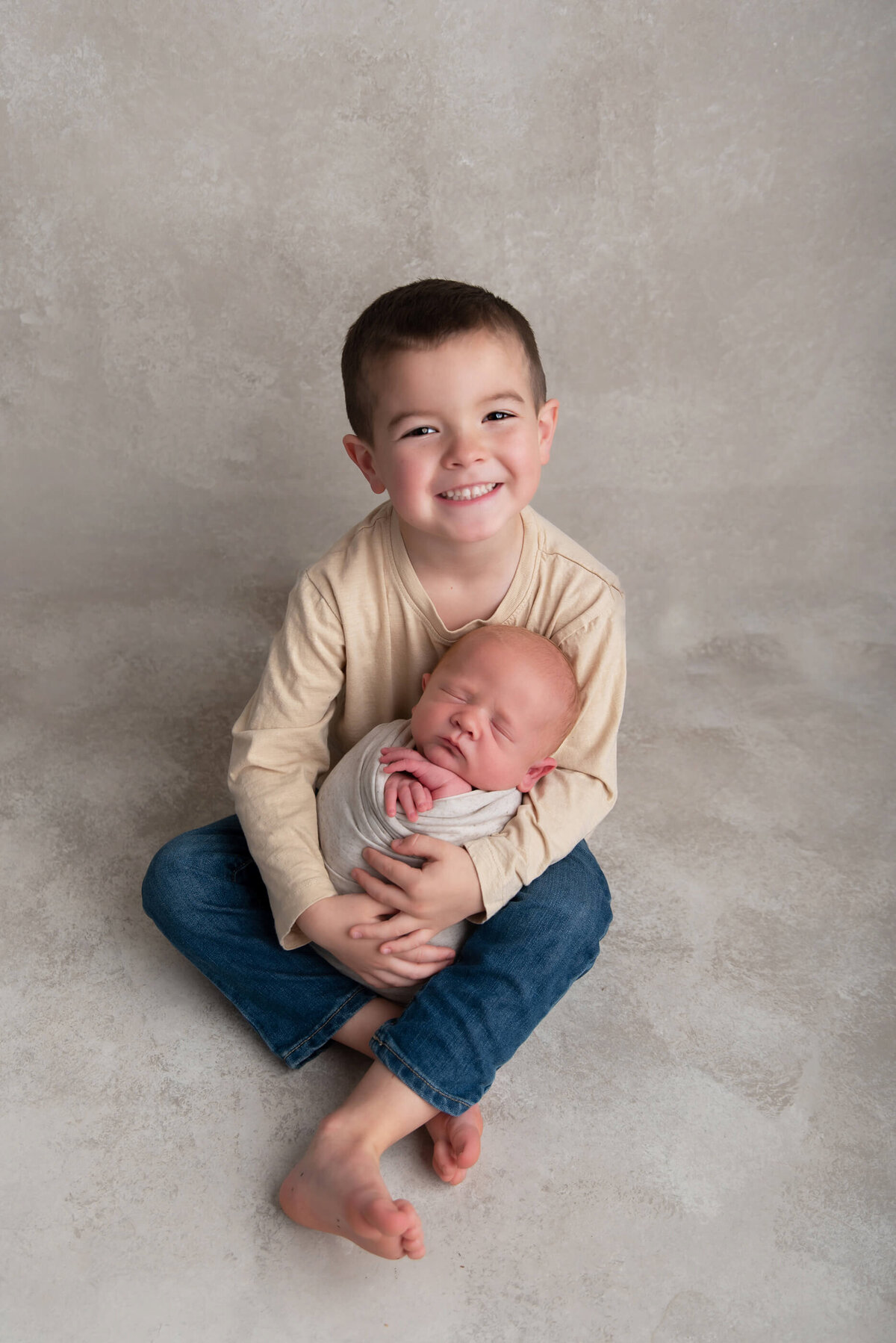 Big brother holding his newborn baby brother on a beige background for the newborn photography session in Burlington, Ontario
