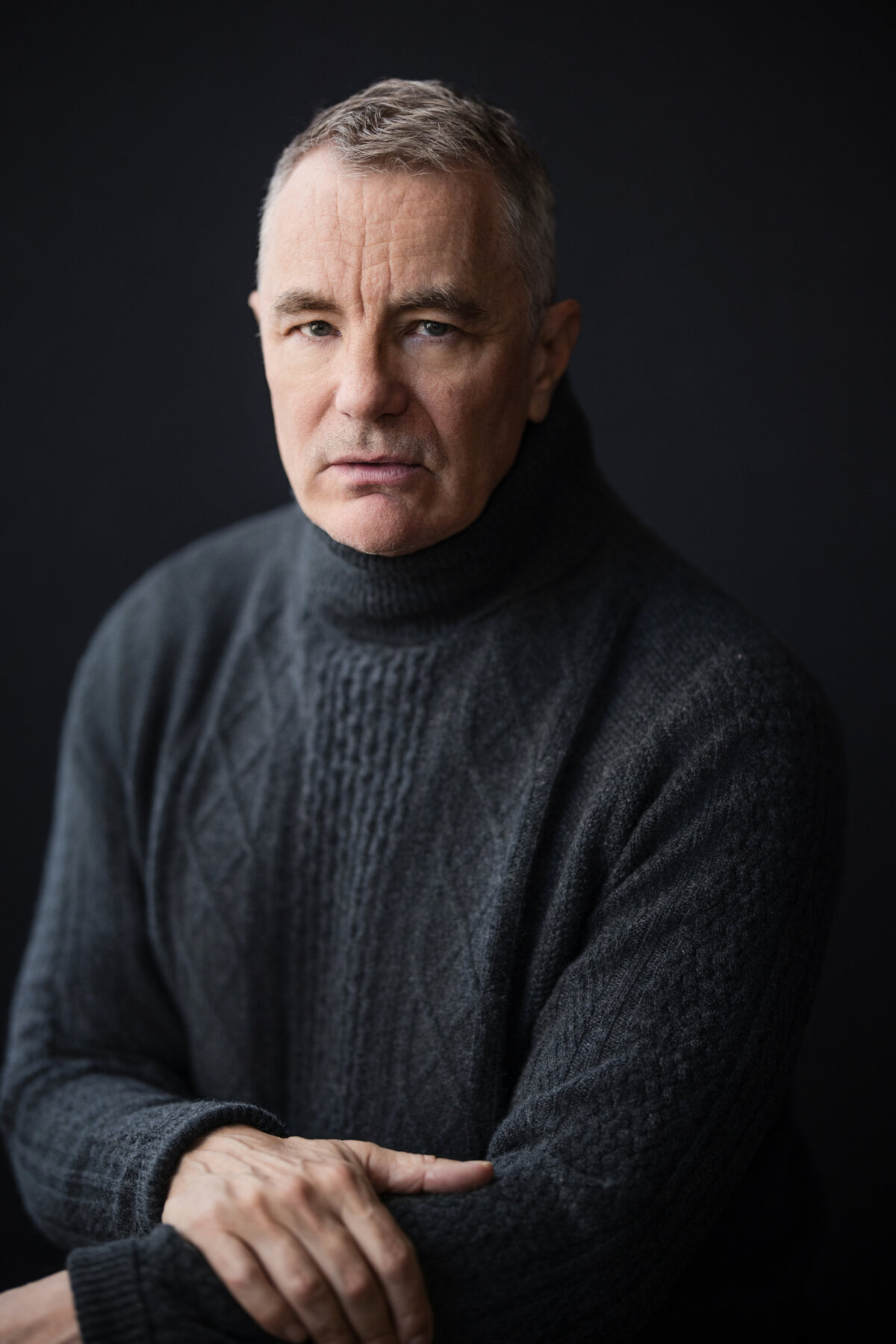 Person seated against a dark background, wearing a textured charcoal turtleneck sweater. They have short, gray hair and a serious expression, looking directly into the camera. The lighting is soft and directional, creating a classic and contemplative headshot.
