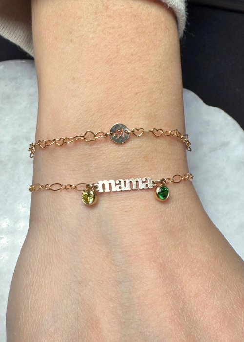 Two custom gold bracelets on a wrist—one with a heart-shaped chain and engraved initial, and another reading "mama" with two green birthstone charms.