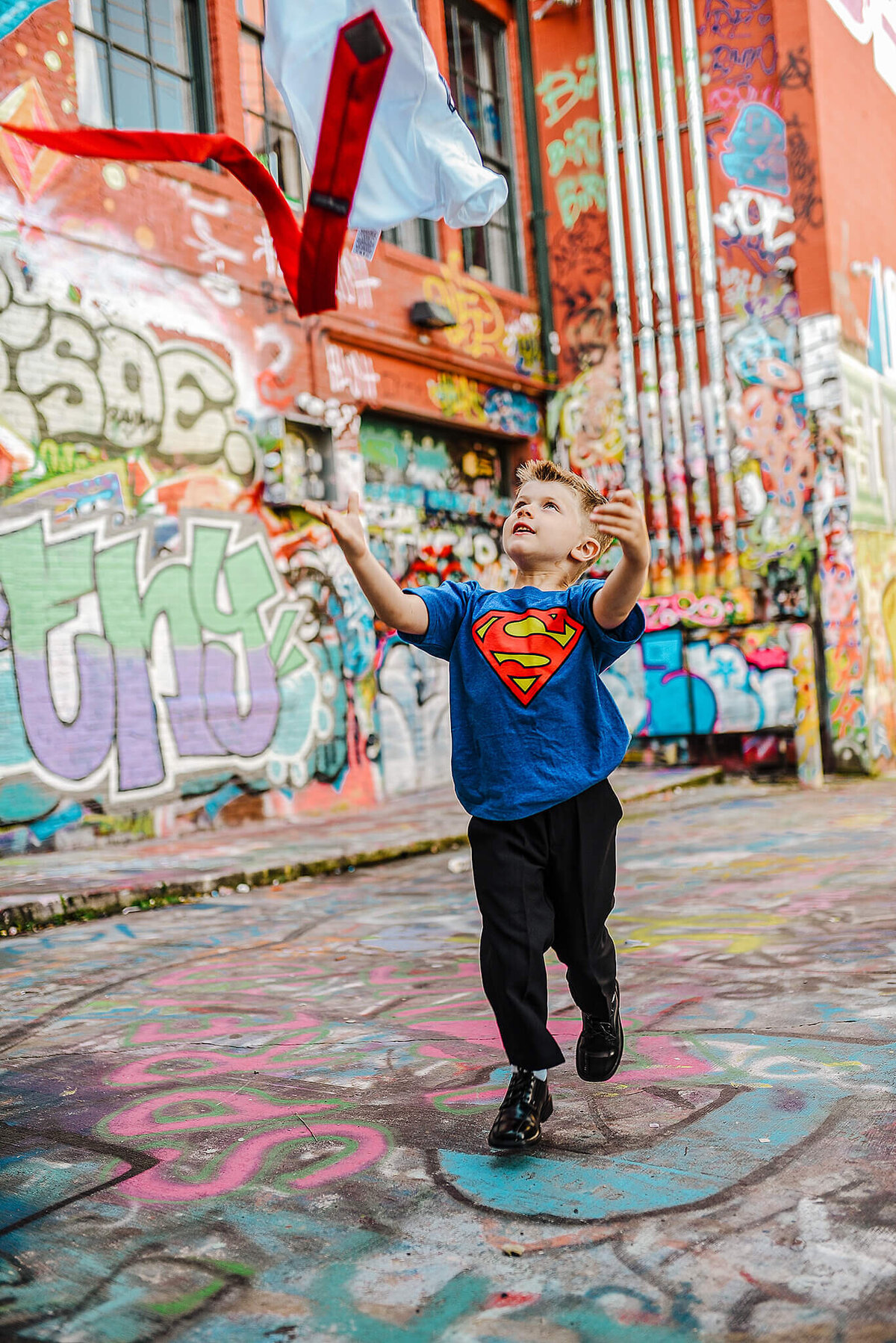 Little boy wearing a superman shirt and black pants throwing a white shirt and red tie into the air in graffiti alley near MICA in Baltimore Maryland