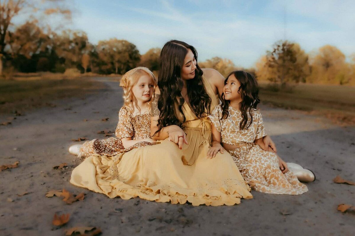 A woman sits on a dirt path outdoors with two young girls, all in long, patterned yellow dresses, surrounded by fallen leaves and trees captured beautifully by a photographer in Warner Robins GA.