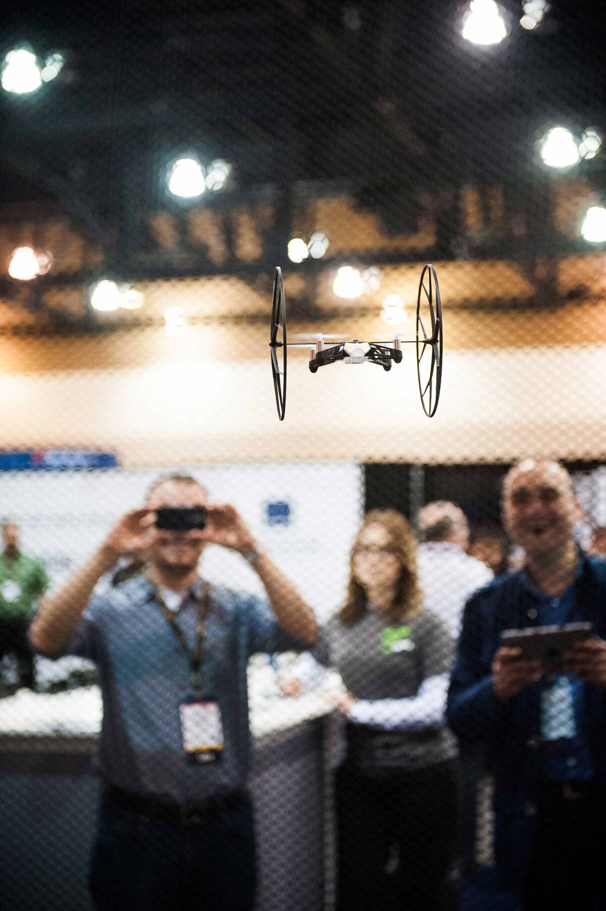 A small drone being flown indoors at engineering conference as onlooker takes a photo in the background.