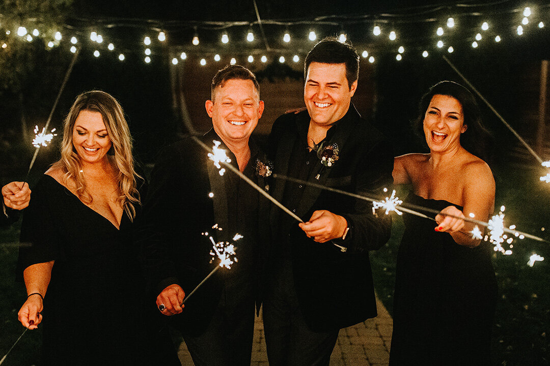 Two grooms wearing black tuxedos hold sparklers while smiling next to two women wearing black gowns.